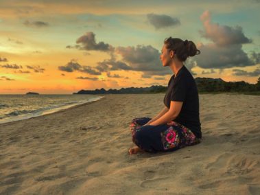 Woman sitting on beach sand doing yoga meditating and relaxing at sunset time